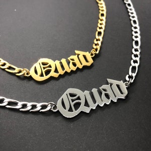 K&ouml;nnte beinhalten: Zwei Halsketten, eine goldene und eine silberne, mit dem Wort "Quad" in altenglischen Lettern.