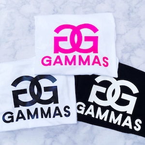 May include: Three white t-shirts with a black and white graphic of the letters "GG" and the word "GAMMAS" printed on them. One shirt has a pink graphic of the letters "GG" and the word "GAMMAS" printed on it.