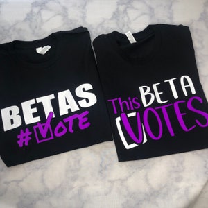 Könnte beinhalten: Zwei schwarze T-Shirts mit weißem und violettem Text. Das erste Shirt sagt "BETAS #VOTE" mit einem violetten Häkchen im Kästchen. Das zweite Shirt sagt "This BETA VOTES" mit einem violetten Häkchen im Kästchen.