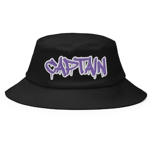 May include: Black bucket hat with the word "CAPTAIN" embroidered in purple with a white outline. The hat has a wide brim and a classic, casual style. This hat is a stylish accessory for everyday wear.