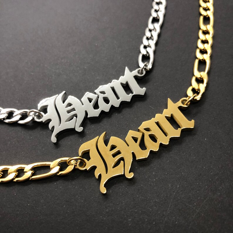 K&ouml;nnte beinhalten: Zwei goldene und silberne Halsketten mit dem Wort "Heart" in gotischer Schrift. Die Halsketten liegen auf einem dunklen Hintergrund.