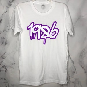 Könnte beinhalten: Weißes T-Shirt mit einer violetten Umrandung des Jahres 1986 in einer Graffiti-Schriftart.