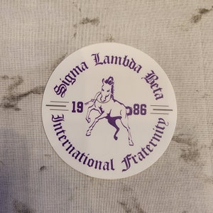 May include: A white circular sticker with purple text and a purple horse illustration. The text reads "Sigma Lambda Beta International Fraternity 1986".
