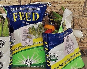 May include: Three reusable tote bags filled with fresh produce and feed bags. The bags are made from recycled feed sacks, featuring blue, white, and green colors. The bags are filled with vegetables and fruits, and the feed bags have text that reads "Certified Organic Feed".