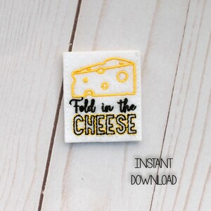 Peut inclure: Patch en feutre blanc avec une broderie noire qui dit "Fold in the CHEESE" et une image brodée jaune d'un bloc de fromage.