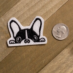 May include: A white felt patch with an embroidered black and white image of a French bulldog peeking over the top of a surface. The bulldog has large, round eyes and a black nose.