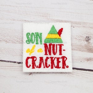 Könnte beinhalten: Weißer Filz-Patch mit rotem Sticktext, der "Son of a Nut-Cracker" lautet, und einer grünen Elfenmütze mit rotem Besatz.