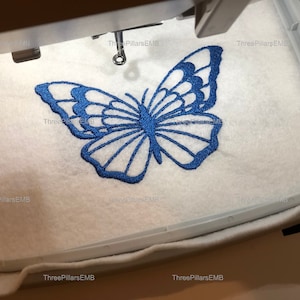 Op de afbeelding: Een blauw vlinderborduurmotief op witte stof in een borduurring.