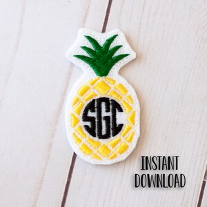 May include: A white felt pineapple with a yellow and black embroidered design. The pineapple has a green felt top and a black monogram in the centre.