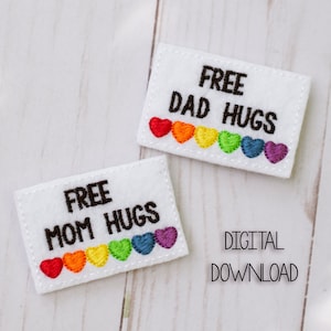 Può includere: Due toppe di feltro bianco con cuori arcobaleno e il testo "FREE MOM HUGS" e "FREE DAD HUGS". Il testo è in ricamo nero. Le toppe sono su una superficie di legno bianco. DIGITAL DOWNLOAD è scritto in nero sotto le toppe.