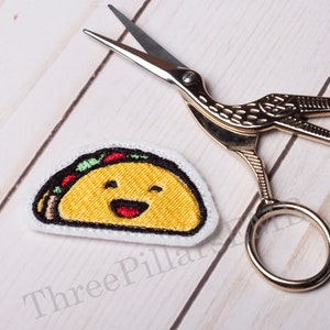 May include: A white felt patch with an embroidered yellow taco with a smiling face. The taco has red and green fillings. The patch is next to a pair of gold scissors.