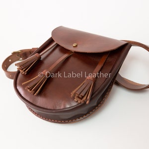 May include: A brown leather satchel with a curved flap and a belt strap. The bag features two decorative tassels and a brass-colored button closure. The bag is approximately 10 inches wide.