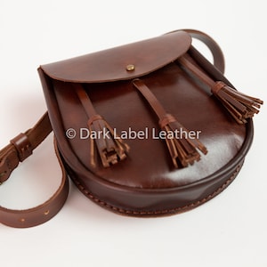 May include: A brown leather shoulder bag with a flap closure and adjustable strap. The bag features decorative tassels and a buckle. The text "Dark Label Leather" is visible on the bag.