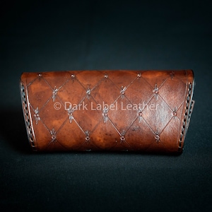 May include: A brown leather wallet with a diamond-shaped pattern and a stitched border. The wallet has the text "Dark Label Leather" embossed on the front.