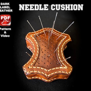 Könnte beinhalten: Ein braunes Ledernadelkissen mit einem genähten Rand und Löchern für Nadeln. Das Kissen ist mit einem weichen Material gefüllt, um die Nadeln zu schützen. Das Kissen ist mit "NEEDLE CUSHION" in weißer Schrift beschriftet.