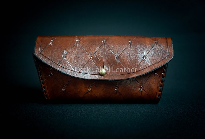 May include: A brown leather glasses case with a quilted design and a gold button closure. The case has a stitched border and the text "Dark Label Leather" is embossed on the front.