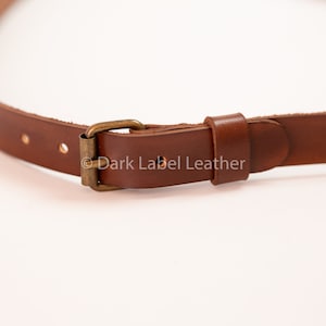 May include: Close-up of a brown leather strap with a brass buckle. The strap has several holes for adjustment and is designed for use with a bag or other accessory. The leather appears to be high-quality and durable.