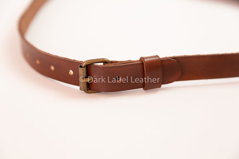May include: Close-up of a brown leather strap with a brass buckle. The strap has several holes for adjustment and is designed for use with a bag or other accessory. The leather appears to be high-quality and durable.