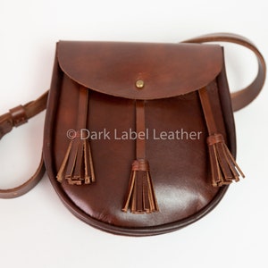 May include: A brown leather shoulder bag with a flap closure and three decorative tassels. The bag has an adjustable strap and a brass buckle. The text "Dark Label Leather" is visible on the bag.