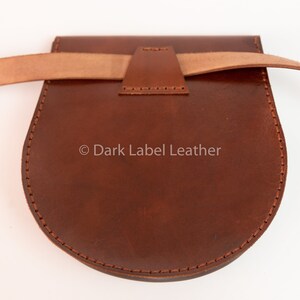 May include: A brown leather pouch with a curved bottom and a flap closure. The pouch has a light brown strap and is stitched with a contrasting thread. The text "Dark Label Leather" is visible on the pouch.