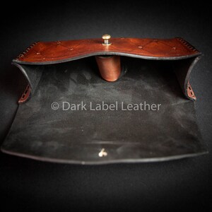 May include: Open brown leather pouch with a black suede interior. The pouch has a decorative stitch pattern and a brass button closure. The text "Dark Label Leather" is visible inside the pouch.