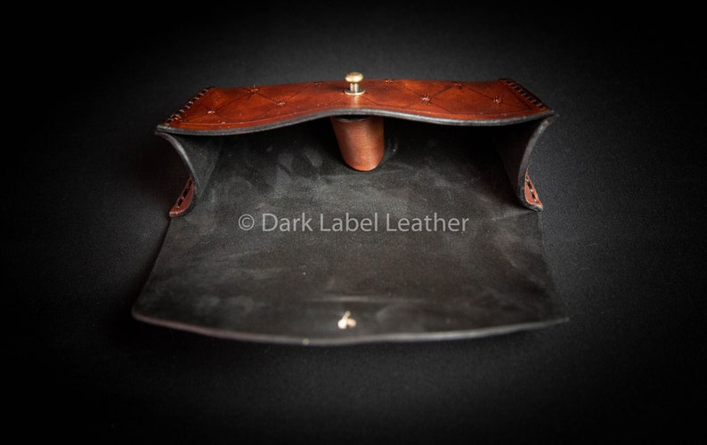 May include: Open brown leather pouch with a black suede interior. The pouch has a decorative stitch pattern and a brass button closure. The text "Dark Label Leather" is visible inside the pouch.