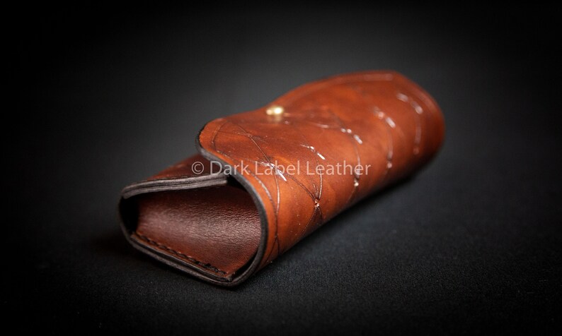 May include: A brown leather case with a decorative pattern and a gold button closure.  &copy; Dark Label Leather