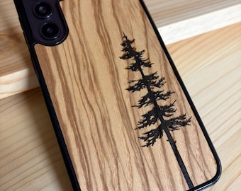 May include: A smartphone case with a light brown wood grain back panel featuring a dark laser-engraved pine tree design. The case has a black frame and a triple camera cutout. The word alto is printed in small black letters at the bottom.