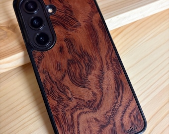 Puede incluir: Una funda para smartphone con incrustación de veta de madera marrón oscuro, marco negro y recorte para triple cámara. La palabra alto está grabada en la esquina inferior derecha del panel de madera, que muestra un patrón de remolinos.