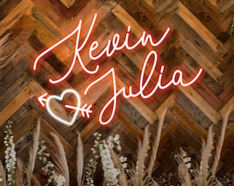 May include: A neon sign with the names "Kevin" and "Julia" in cursive script, with a heart and arrow between the names. The sign is mounted on a wooden wall with a chevron pattern. The sign is surrounded by a variety of dried flowers and grasses.