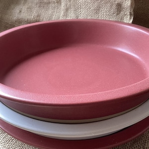 Op de afbeelding: Een stapel van drie keramische serveerschalen. De bovenste schaal is een gedempt rood, de middelste schaal is wit en de onderste schaal is ook gedempt rood. De schalen zijn rond, ondiep en hebben een licht verhoogde rand. De achtergrond is een textiel, bruinachtig stof.