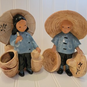 Puede incluir: Dos figuras de cerámica que representan figuras con vestimenta tradicional. Llevan grandes sombreros de paja y camisas azules. Uno sostiene un pergamino, el otro una cesta. Las figuras son de color beige, azul, negro y blanco.