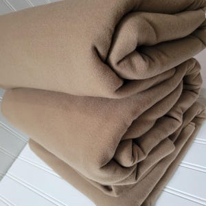 Op de afbeelding: Een stapel van drie gevouwen dekens in een warme bruintint. De dekens zijn netjes gerangschikt en tonen hun zachte textuur en de plooien die door hun rangschikking zijn ontstaan. De dekens liggen op een wit oppervlak met verticale lijnen.