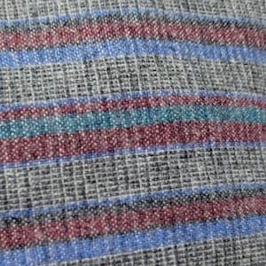 May include: Close-up of a woven fabric with horizontal stripes. The fabric has a gray base with stripes of blue, teal, and burgundy. The texture appears slightly rough, with visible threads creating a grid-like pattern.