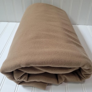 Op de afbeelding: Een gevouwen, bruin gekleurde stof, mogelijk een deken of sprei, staat centraal. De stof is zacht en lijkt gemaakt te zijn van een zacht materiaal. De vouwen creëren een gelaagd effect en de algehele presentatie is netjes.