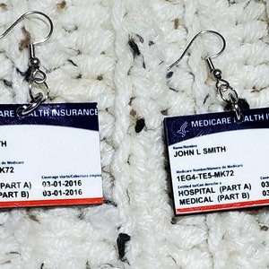 May include: A pair of earrings featuring miniature replicas of Medicare health insurance cards. The cards are white with blue and red accents and feature the text "MEDICARE HEALTH INSURANCE", "JOHN L SMITH", "1EG4-TE5-MK72", "HOSPITAL (PART A)", "MEDICAL (PART B)", "Coverage Starts/Cobertura empieza", "03-01-2016".