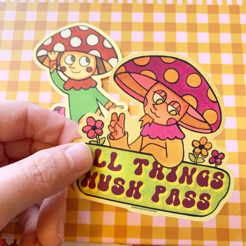 May include: Two cartoon mushroom characters with smiling faces and peace signs. The larger mushroom is yellow with a pink cap and says "All Things Mush Pass".