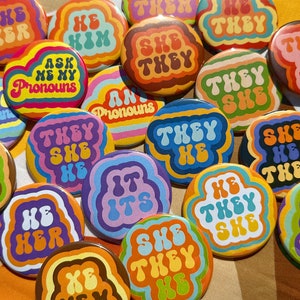 May include: A collection of colorful round buttons with various pronoun combinations in a retro font style. The buttons feature phrases like "She Her", "He Him", "They She He", and "Ask Me My Pronouns".