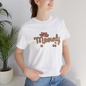 May include: White t-shirt featuring the word "Meowdy" in a brown, Western-style font, with a cowboy hat, paw print, and cherries graphic. The short-sleeved shirt is made of a soft material.