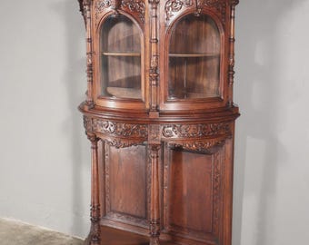 May include: A dark brown, antique corner cabinet with ornate carvings. The cabinet features two arched glass doors on top and solid wood panels below. The piece has a curved design and sits on a triangular base, showcasing a classic, vintage style.