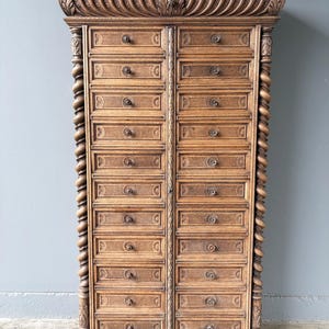 Puede incluir: Un armario alto de madera con dos columnas verticales de doce cajones cada una. La pieza est&aacute; adornada con columnas talladas en espiral a ambos lados y una cornisa decorativa tallada en la parte superior. Cada caj&oacute;n tiene un peque&ntilde;o pomo redondo.