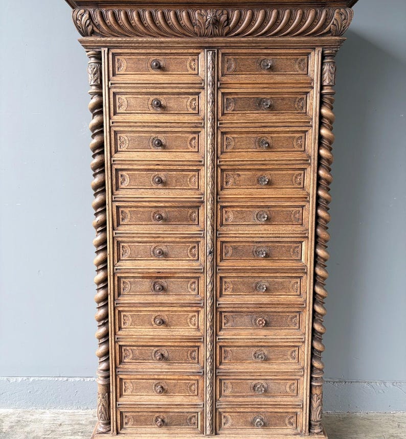 Puede incluir: Un armario alto de madera con dos columnas verticales de doce cajones cada una. La pieza est&aacute; adornada con columnas talladas en espiral a ambos lados y una cornisa decorativa tallada en la parte superior. Cada caj&oacute;n tiene un peque&ntilde;o pomo redondo.