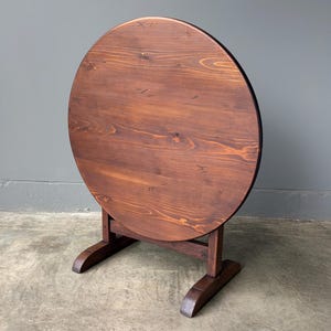 May include: A round wooden table with a dark brown stained finish and visible wood grain patterns. The table features a circular top supported by a sturdy base with two horizontal feet, designed to stand upright in a vertical position.