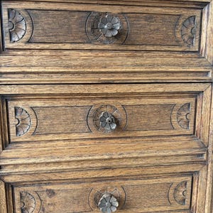 Puede incluir: Una c&oacute;moda de madera marr&oacute;n con tres cajones horizontales. Cada frente de caj&oacute;n est&aacute; decorado con patrones semicirculares tallados en los lados y un pomo floral circular central. La veta de la madera es visible en toda la pieza.