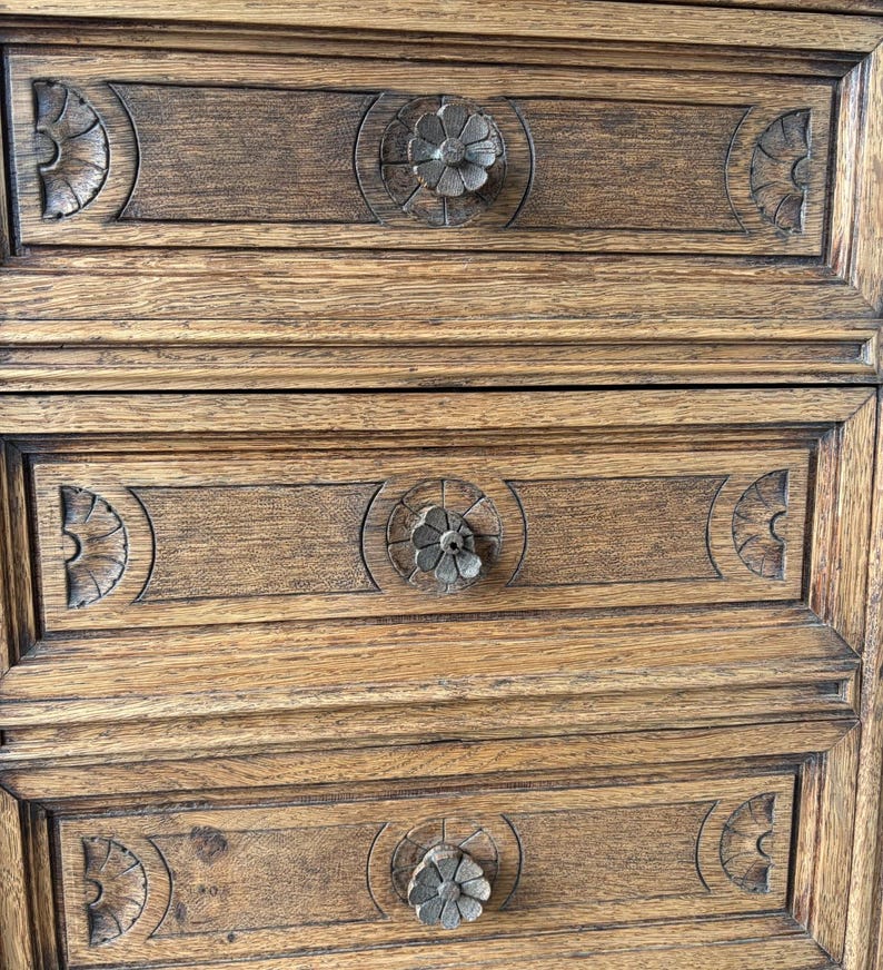 Puede incluir: Una c&oacute;moda de madera marr&oacute;n con tres cajones horizontales. Cada frente de caj&oacute;n est&aacute; decorado con patrones semicirculares tallados en los lados y un pomo floral circular central. La veta de la madera es visible en toda la pieza.