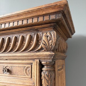 Puede incluir: Un mueble de madera marr&oacute;n claro con intrincados detalles tallados, que incluyen un friso decorativo con ranuras verticales, una cornisa con motivos de hojas y un elemento de columna tallado en el lateral. Se ve un pomo redondo de madera.