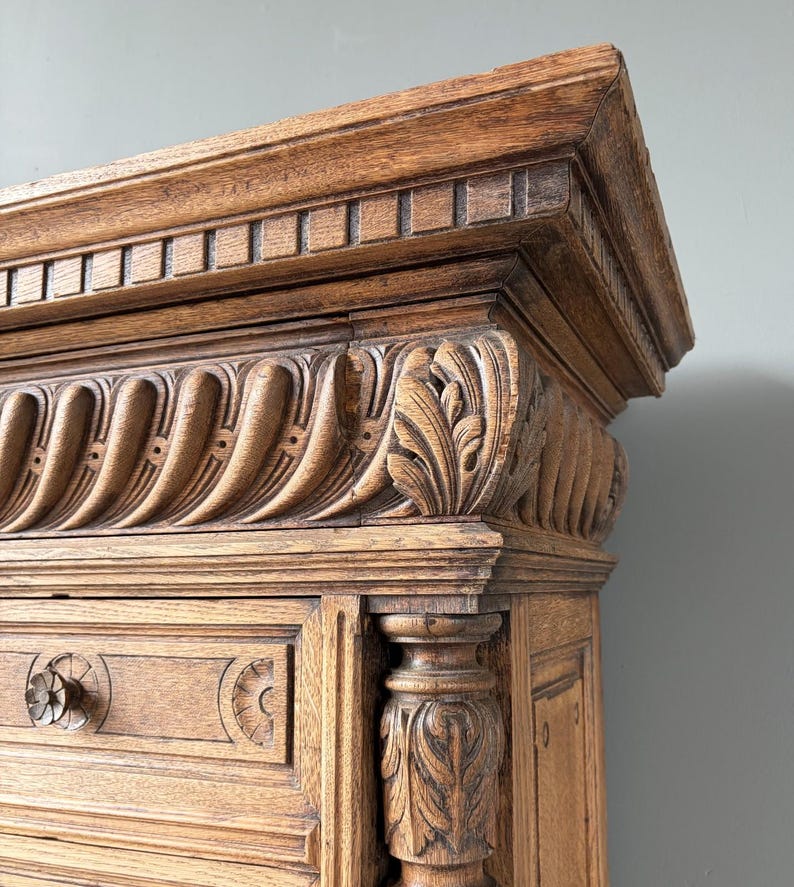 Puede incluir: Un mueble de madera marr&oacute;n claro con intrincados detalles tallados, que incluyen un friso decorativo con ranuras verticales, una cornisa con motivos de hojas y un elemento de columna tallado en el lateral. Se ve un pomo redondo de madera.