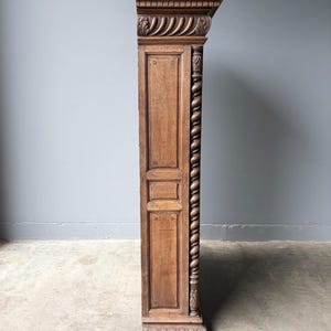 Puede incluir: Un panel lateral de armario de madera alto y estrecho con un acabado marr&oacute;n medio. La estructura incluye una cornisa superior tallada, un detalle de columna vertical retorcida y paneles rectangulares empotrados con pasadores visibles.