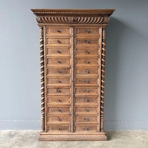 Puede incluir: Un armario rectangular alto de madera de color marr&oacute;n medio con dos columnas verticales de doce cajones cada una. La pieza presenta detalles tallados intrincados, incluyendo columnas retorcidas en los laterales y una cornisa decorativa.