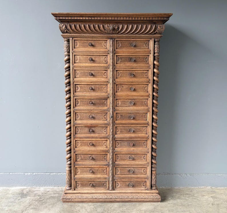 Puede incluir: Un armario rectangular alto de madera de color marr&oacute;n medio con dos columnas verticales de doce cajones cada una. La pieza presenta detalles tallados intrincados, incluyendo columnas retorcidas en los laterales y una cornisa decorativa.